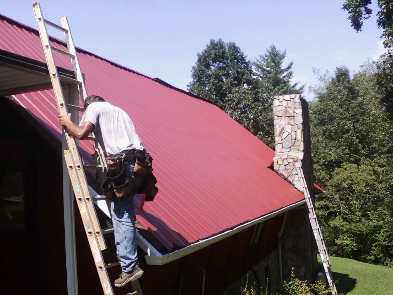 Roofer on a ladder beside a red metal roof and stone chimney — Soffit & Fascia Repair by Roofing Brothers in Moorefield